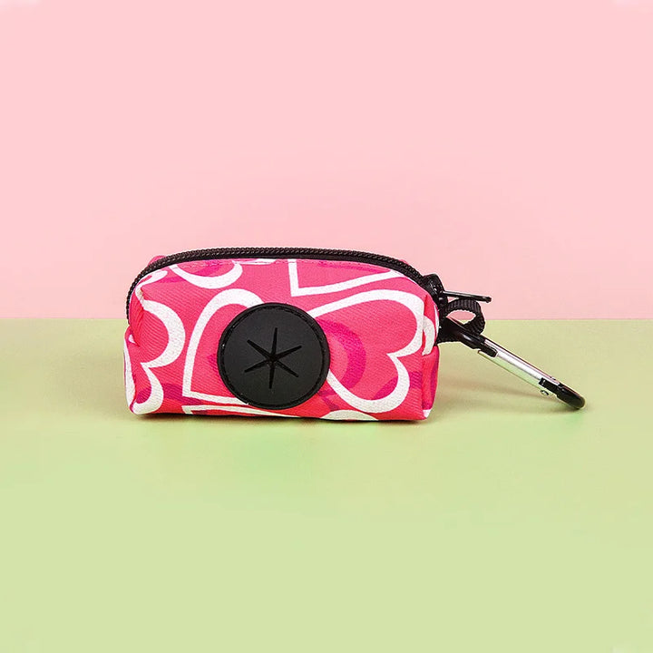 Pink heart-patterned Dog Poop Bag Holder with a black zipper and keychain on a pink and green gradient background